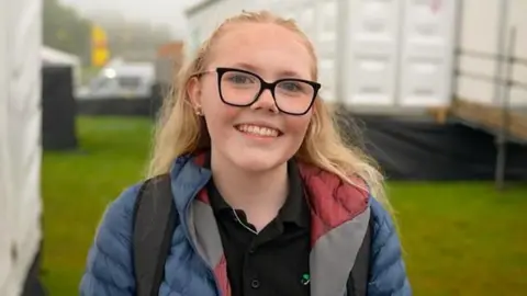 Cari is smiling at the camera. She is wearing glasses and her long blonde hair is pulled away from her face, She is wearing a blue puffa coat with a black polo shirt, with grass behind her. 