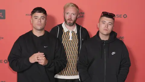 PA Media Kneecap members Móglaí Bap, DJ Próvaí and Mo Chara wearing dark clothing and standing against a red background with black writing on it.
