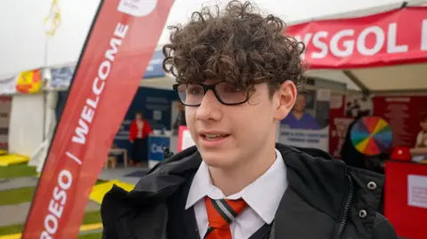 Hari looking to the side. He is wearing glasses and has brown curly hair. He is wearing a white shirt, red tie, black jumper and black coat and standing in front of a red stall. 
