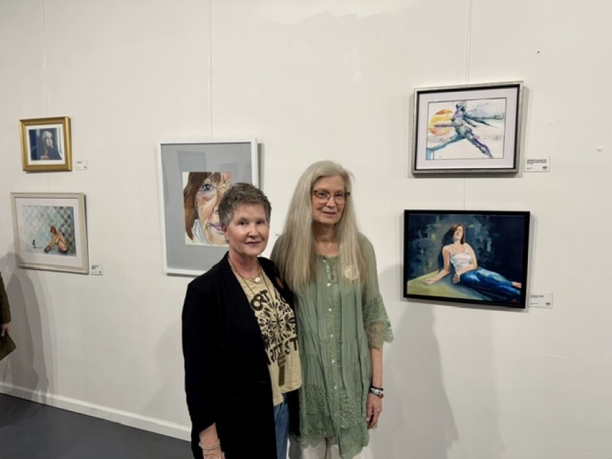 Two women standing and smiling in front of a white wall displaying several framed paintings, including portraits and animal artwork, in an art gallery setting.