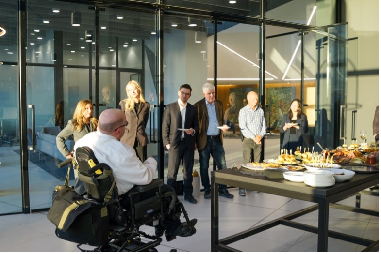 A man in an electric wheelchair and dress shirt addresses a group of people in a corporate board room of glass windows and natural evening lighting.