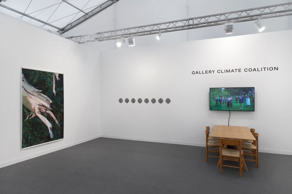 Installation view of the Gallery Climate Coalition booth with a large photograph of a splintered tree trunk on the left wall, a horizontal row of small circular artworks on the center wall, a video screen showing a group standing on grass, and a wooden table with chairs beneath the screen. The setting is a white-walled art fair tent.