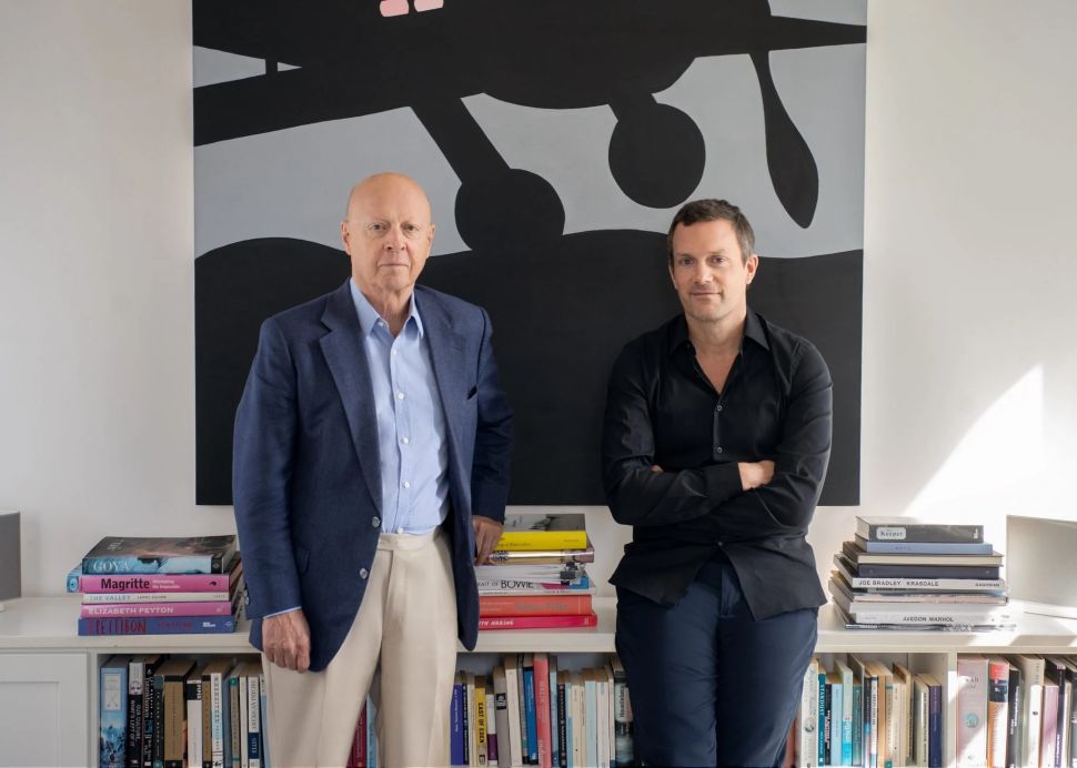 Two men stand in front of a black-and-grey abstract painting, surrounded by art books; one wears a blue blazer and beige trousers, the other a black shirt and dark pants.