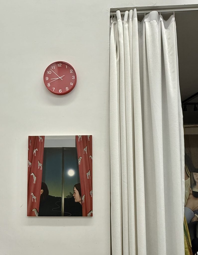 A painting depicting a bright full moon seen from a window with red curtains on both sides of the window hung on a white wall. Above it is a red clock. On the right is a large white curtain covering a storage space for paintings.