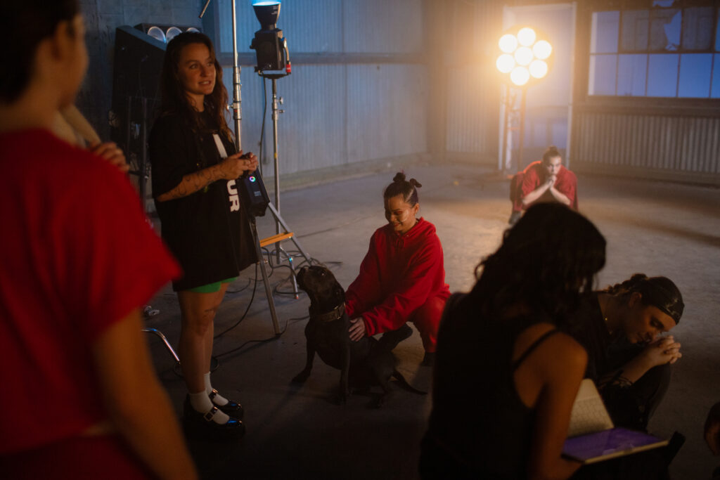 A female choreographer addressing dancer on set. One dancer pets a dog on the floor.