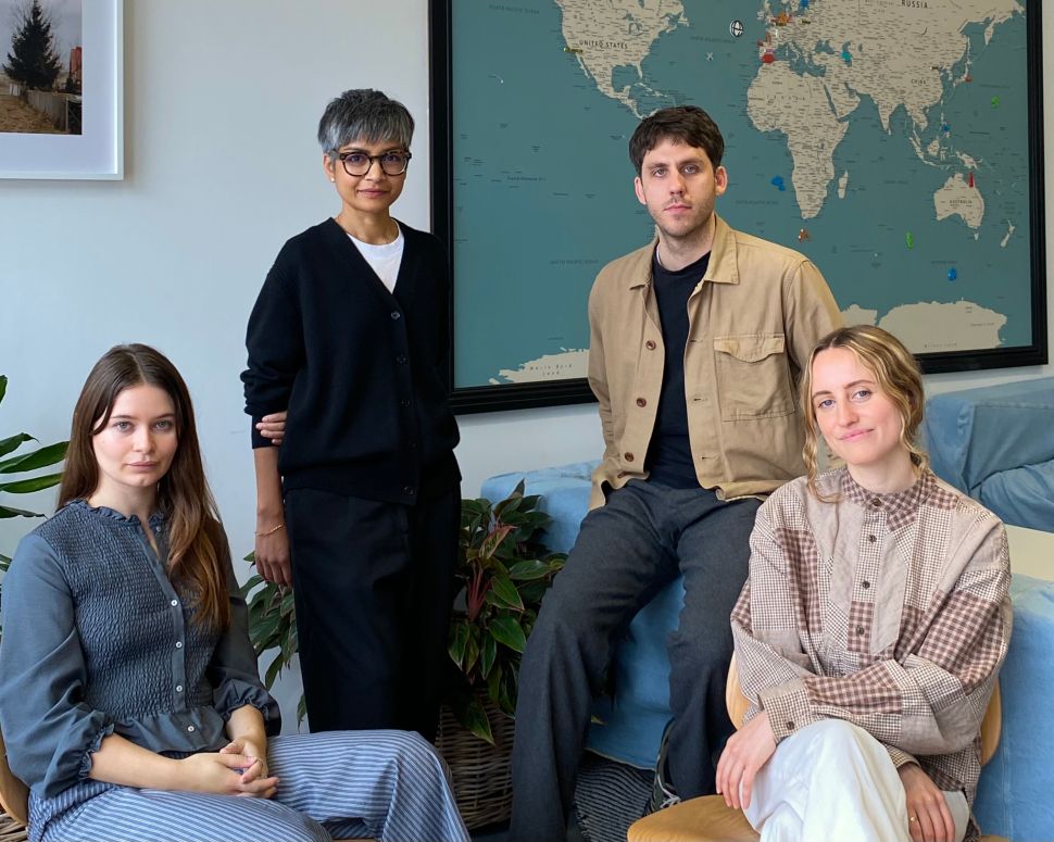 A group portrait of four people seated and standing in an office environment in front of a large world map. Two women sit in front, while a woman and a man stand behind them. A framed photograph and houseplants are visible in the background.