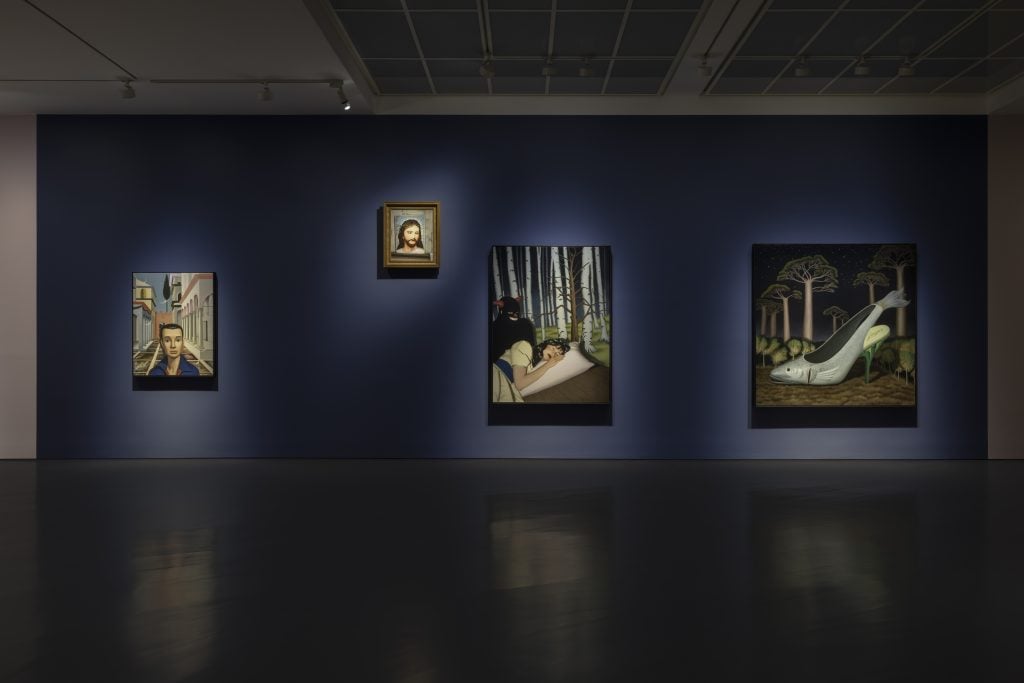 Four paintings of various sizes hung on a navy blue wall in a dimly lit gallery space