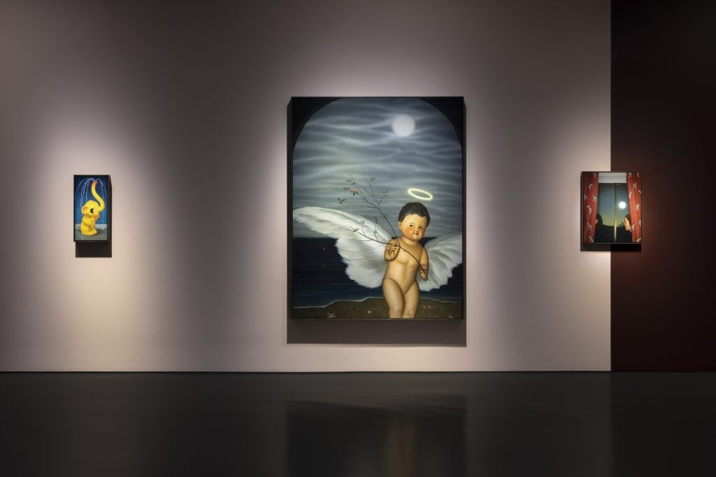 Three paintings, including one large painting depicting an image of a cute angel with wings in the middle, hung on a large wall in a gallery space.