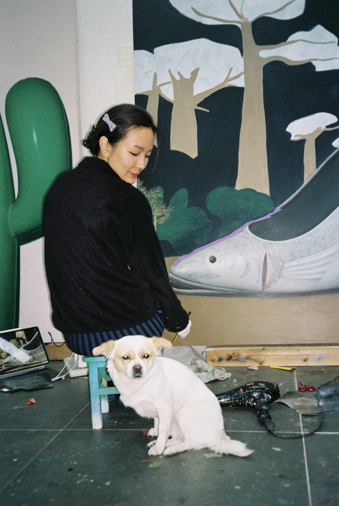 An East Asian woman in black top sitting on a blue stool in front of a painting of a fish. She turns around to look at a dog in beige colour.