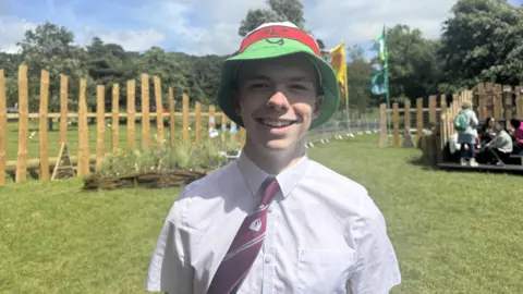 BBC Cai, wearing a white shirt, burgundy tie with red stripes, and a white, red, and green striped bucket hat with Mistar Urdd's face on it. He is standing on the Maes, with grass and wooden fencing behind him. 