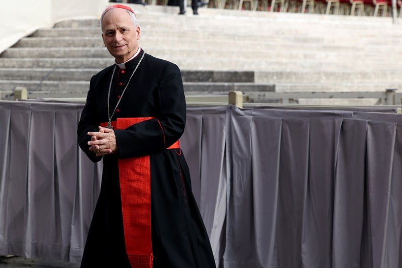 A cardinal in a black robe with red sash with hands folded in front of him walks past a stage and steps.