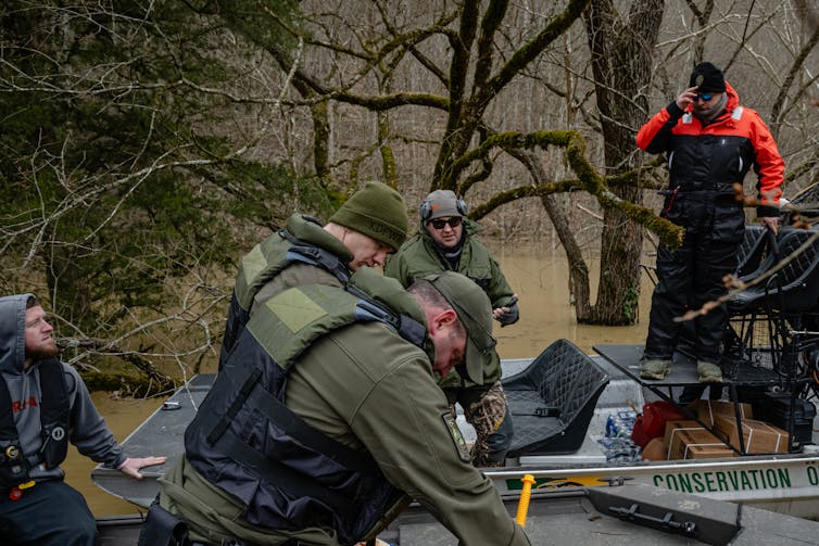a group of men wearing uniforms and life vests stand around boats in muddy water