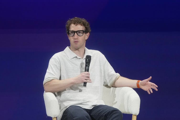 A man wearing a white shirt and big black glasses speaks into a microphone.