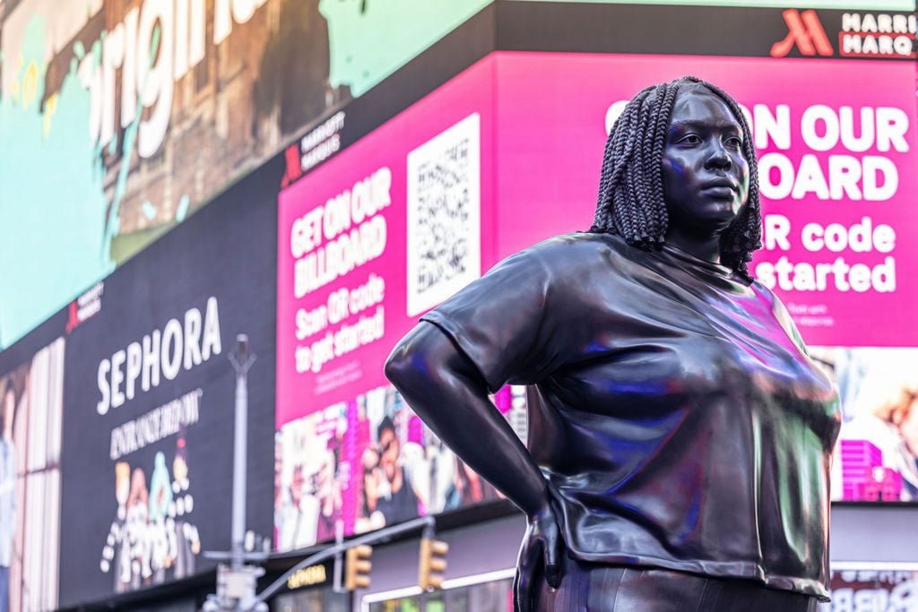 An oversize bronze sculpture of a Black woman with braids, hands on hips, stands amid the advertisements of New York's Times Square