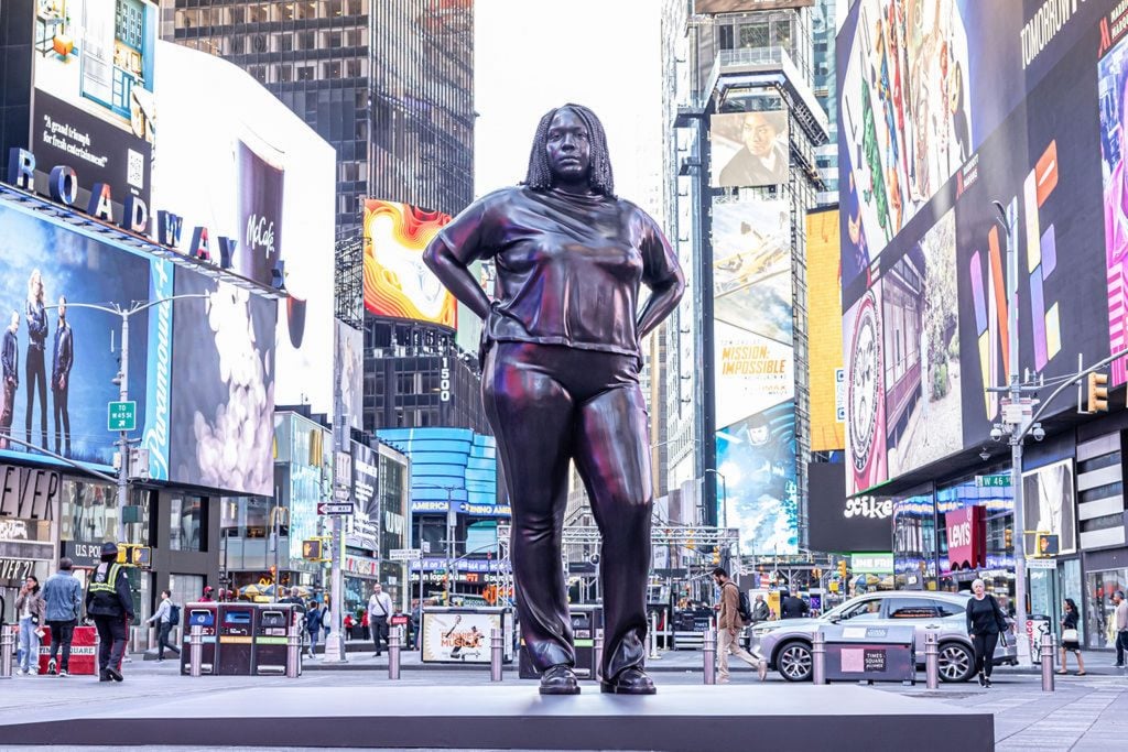 An oversize bronze sculpture of a Black woman with braids, hands on hips, stands amid the advertisements of New York's Times Square
