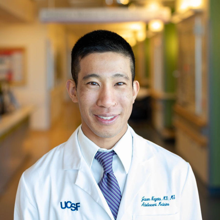 Jason Nagata wears a white doctor's coat and stands in a hostpial hallway