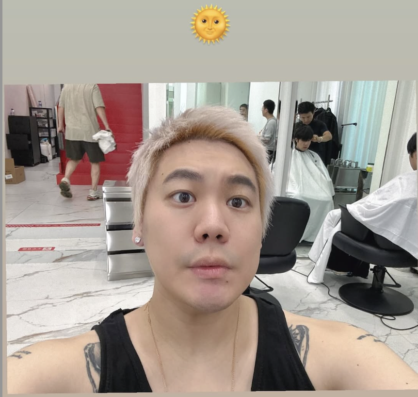 Selfie taken in a hair salon by a person with platinum blonde hair, wearing a black tank top. Behind them, several clients are getting haircuts, and a man in shorts walks by with a towel. The Instagram story caption is a sun emoji.