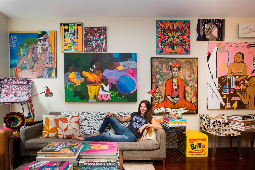 A woman lounges barefoot on a mid-century modern sofa in a vividly decorated living room, surrounded by colorful contemporary artworks. Framed paintings fill the wall behind her, including a large portrait of Frida Kahlo, a vibrant abstract, and a playful depiction of Black figures with balloons. A Brillo soap pad box and art books stacked on the coffee table and floor add to the eclectic, art-filled ambiance. The woman wears a “Rock Girl” t-shirt and jeans, smiling confidently at the camera.
