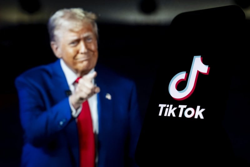 Donald Trump points toward a Tiktok logo on a dark phone screen.