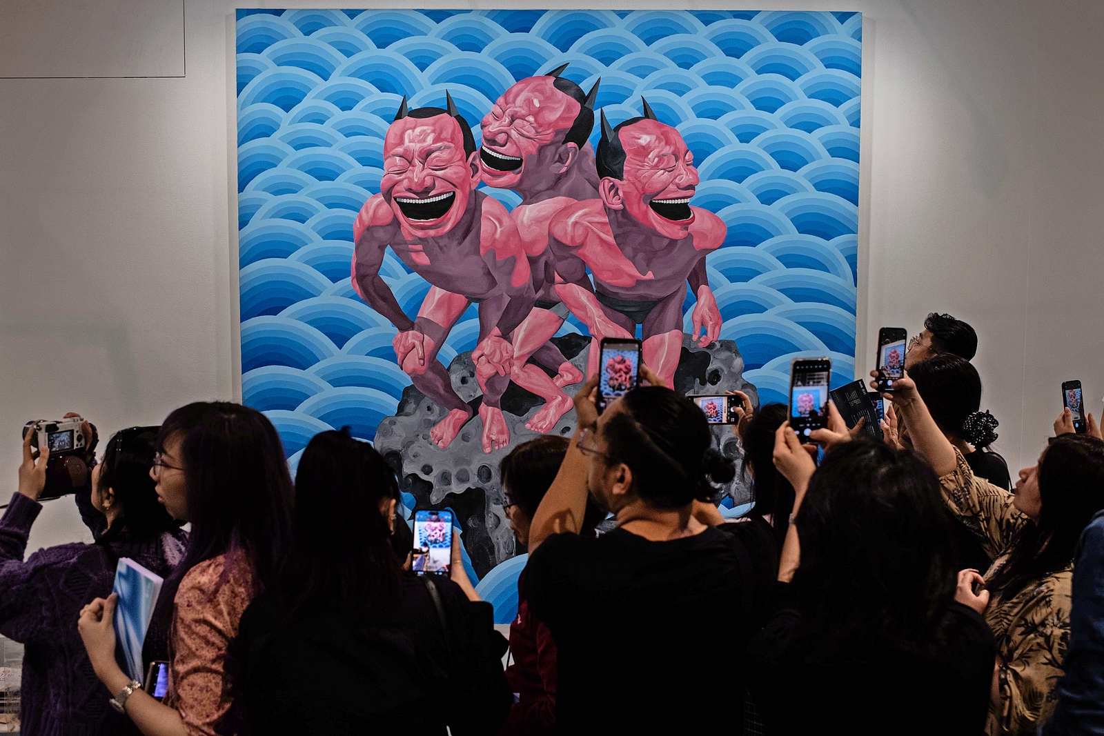 People photographing a painting of three laughing figures on a rock surrounded by blue waves at Art Basel Hong Kong.