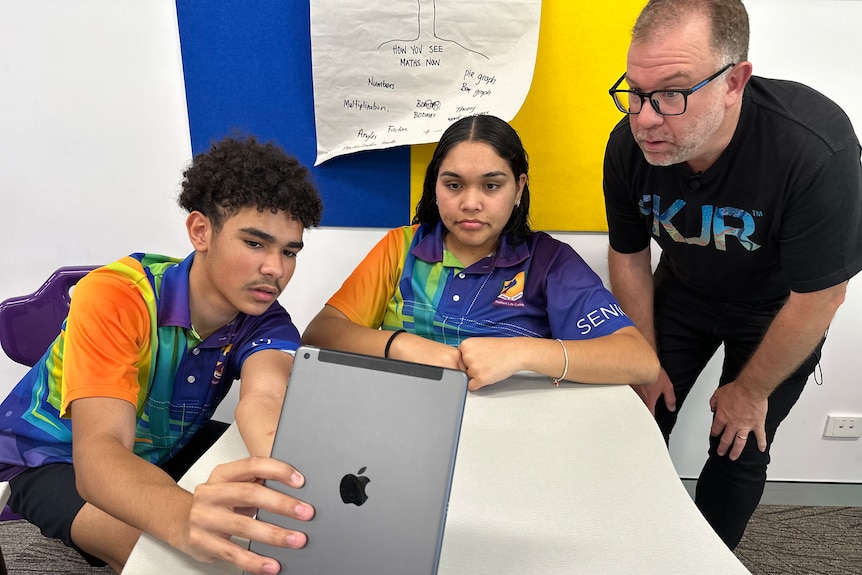 A white man stands over two young teenagers as they play with a tablet in a classroom