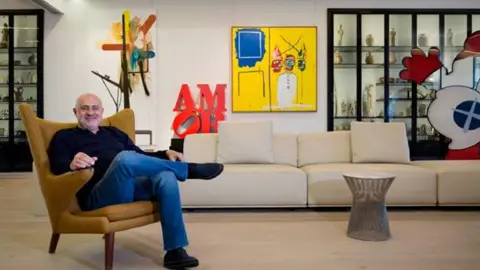 Selection Arts Ahmad sits on an armchair with his legs crossed in his penthouse in Beirut. He is wearing jeans and a dark shirt. Behind him is a wall of art and a cabinet with objects in. There is a cream sofa against the wall.