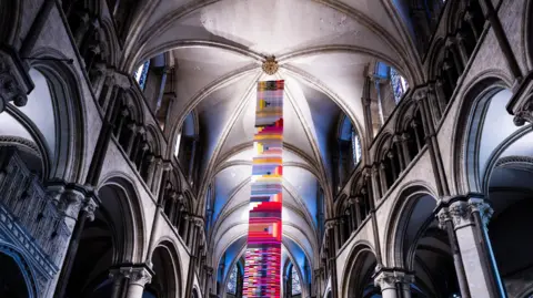 Jo Burn / BBC A long rectangular and colourful tapestry hangs from the ceiling of a cathedral.