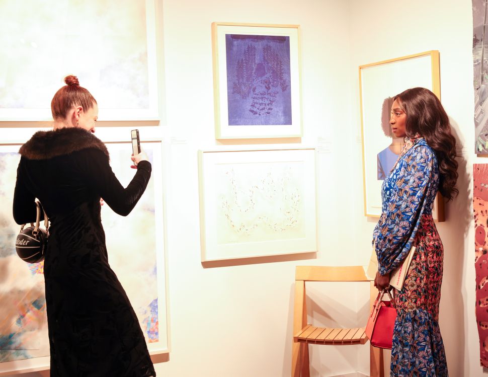 A woman in a long black fur lined jacket takes a picture of another woman wearing a printed dress at a print fair