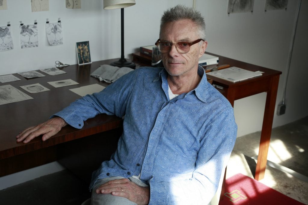 An older man with short gray hair and glasses sits at a wooden desk in a studio space. He wears a light blue patterned button-up shirt and beige trousers. Behind him are drawings pinned to the wall and art materials scattered across the desk. Sunlight streams in from the right, casting soft shadows across the room.