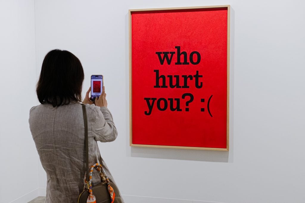 a woman photographs a painting with black text on a red background reading who hurt you?