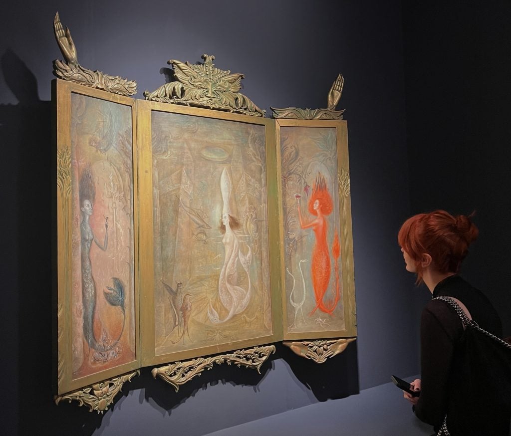 A viewer stands closely examining an ornate triptych painting encased in an elaborate frame with sculpted hands and foliage. The central panel features a pale female figure with long hair and flowing robes; the side panels depict mystical beings in reddish and bluish hues. The piece has a surreal and mythological atmosphere.