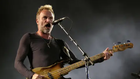 PA Media Sting holding a guitar and singing into a microphone on a stand.