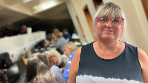 BBC A woman with blonde hair and red-rimmed glasses, and wearing a sleeveless black and white shirt, stands in an office and smiles. People can be seen talking and walking behind her.