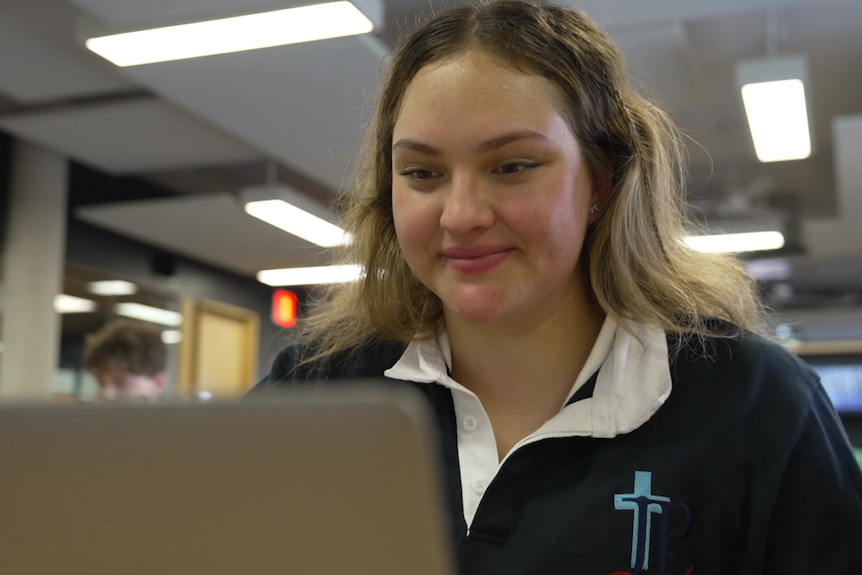 A young white girl with long brown hair looking at a laptop in a classroom and smiling